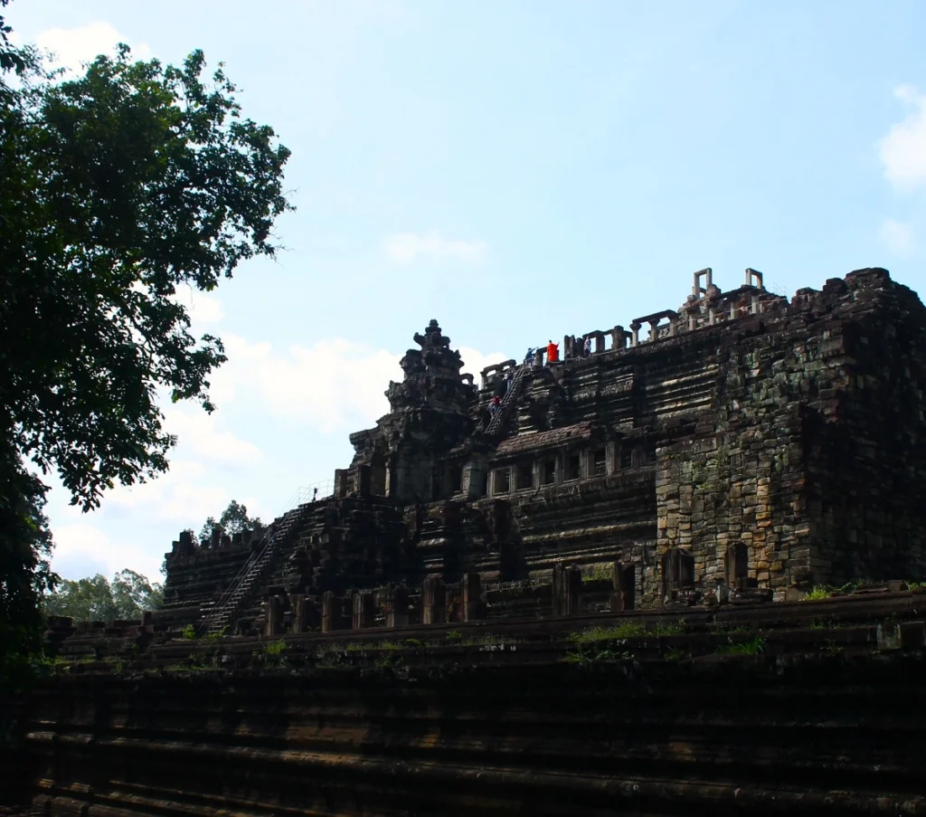 Ansicht der Hauptpyramide des Baphuon-Tempels in Angkor Thom mit einem Mönch in orangefarbenem Gewand auf der Spitze.