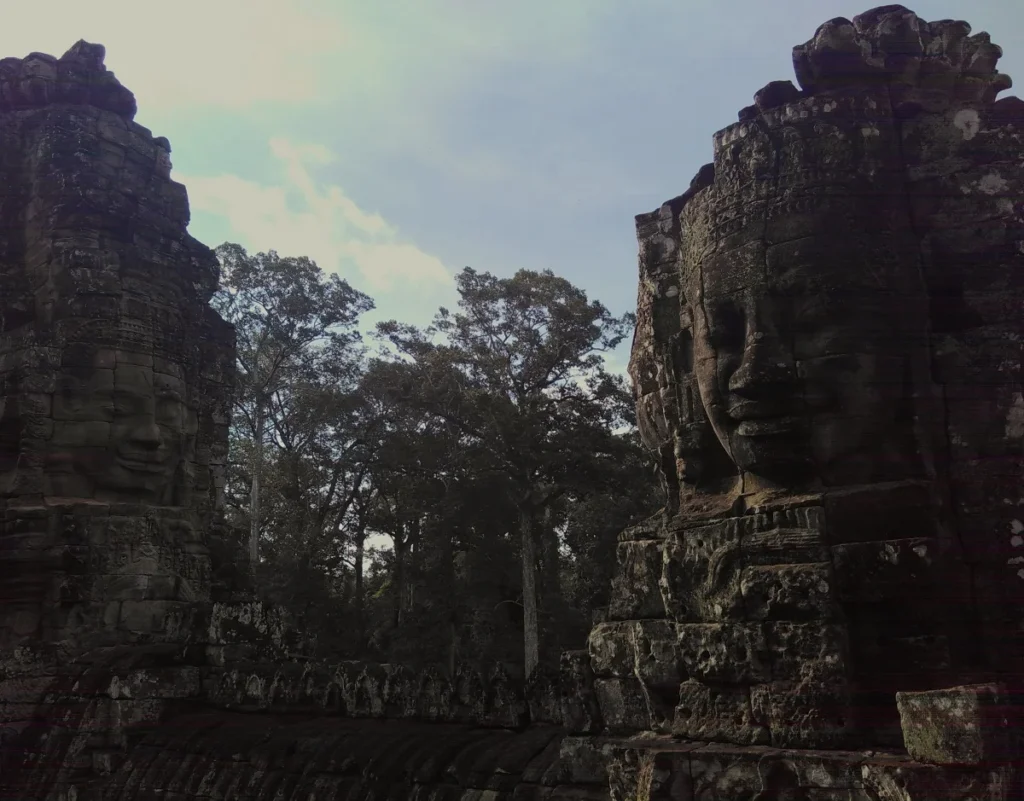Kunst im Khmer-Reich. Nahaufnahme von zwei freundlichen Gesichtern auf zwei Türmen des steinernen Bayon-Tempels. Im Hintergrund Bäume und bewölkter Himmel. 