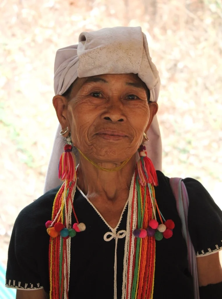 Nahaufnahme einer älteren, lächelnden Karen Frau im Mae Klang Luang Village. Sie trägt ein weißes Kopftuch, bunte Quasten-Ohrringe und eine schwarze Bluse mit weißem Rand. 