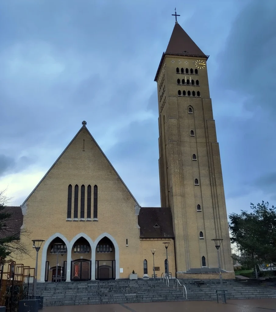 Kirche Sint-Martinus in Genk von leicht unten nach oben fotografiert. Rechts Kirchturm mit Kreuz an Spitze. Links Schiff mit 3 neogotischen Bogen-Eingängen. Treppenaufgang zur Kirche.