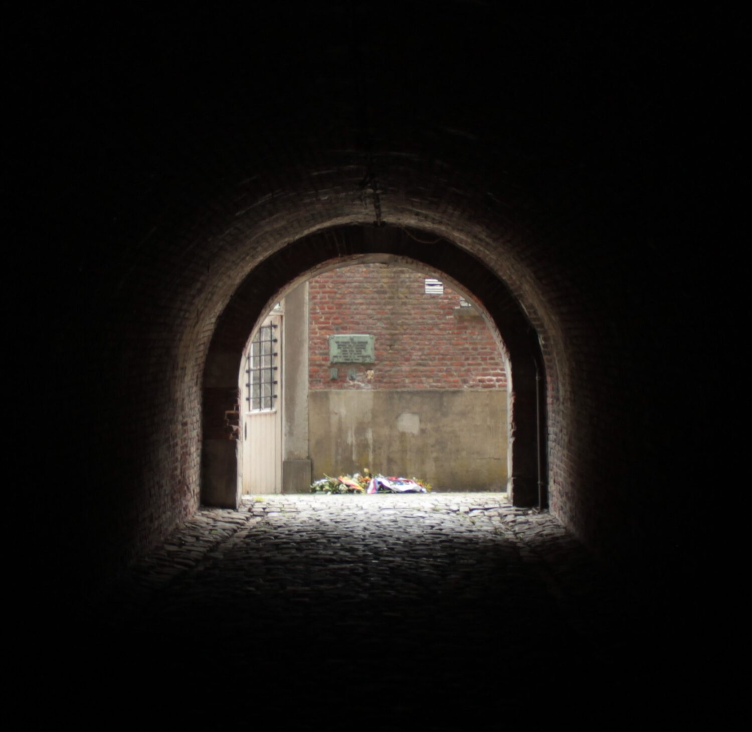 Dunkler Backsteintunnel führt zu hellem Hof mit Gedenkkränzen . 