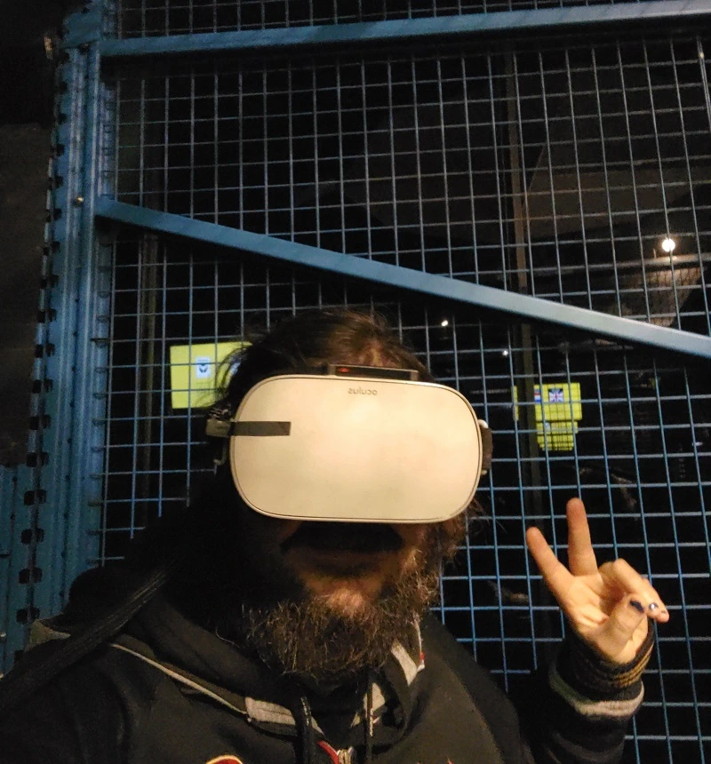 Bärtige Person mit Virtual Reality Brille vor blauem Gitter macht Peace-Zeichen mit linker Hand. 