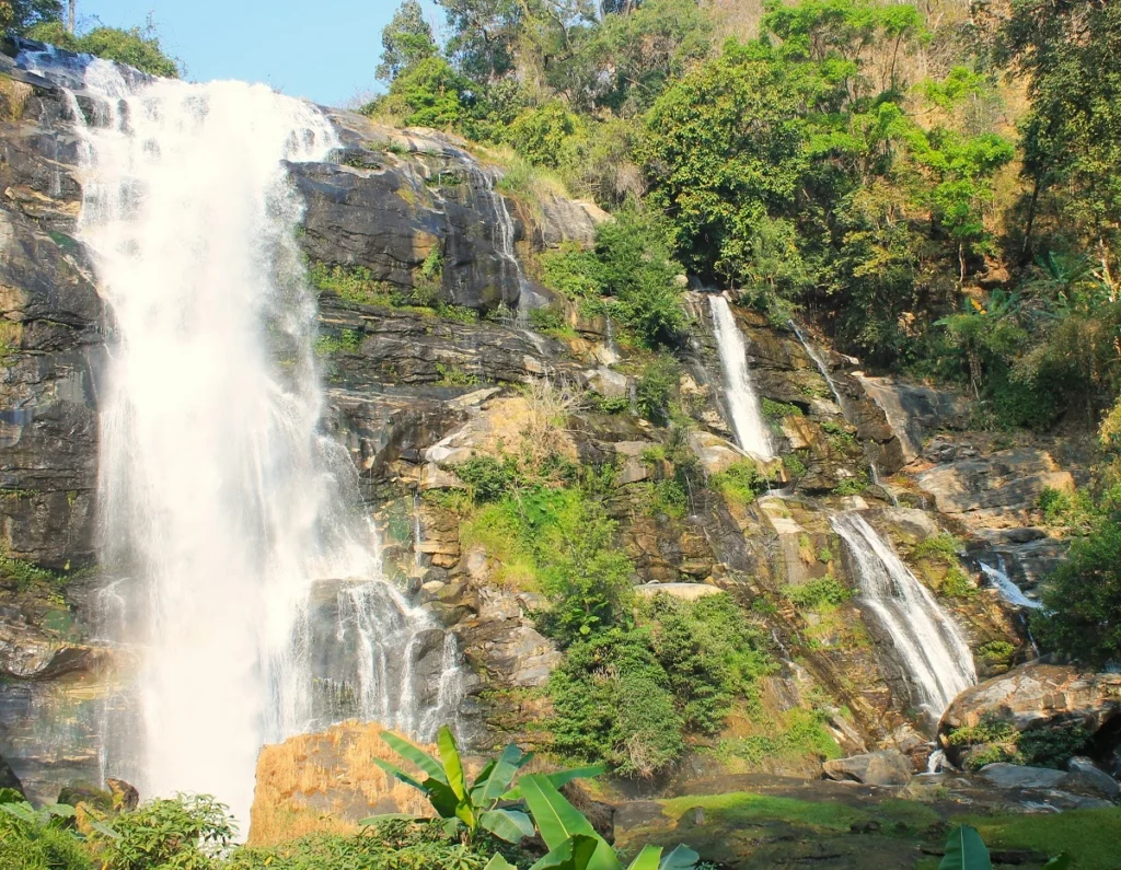 Wasserfall Wachirathan. Kaskadenartige Felswand. Links großer Wasserfall. Rechts kleinere Ströme von fallenden Wasser. Felsen umgeben von grünen Bäumen. 