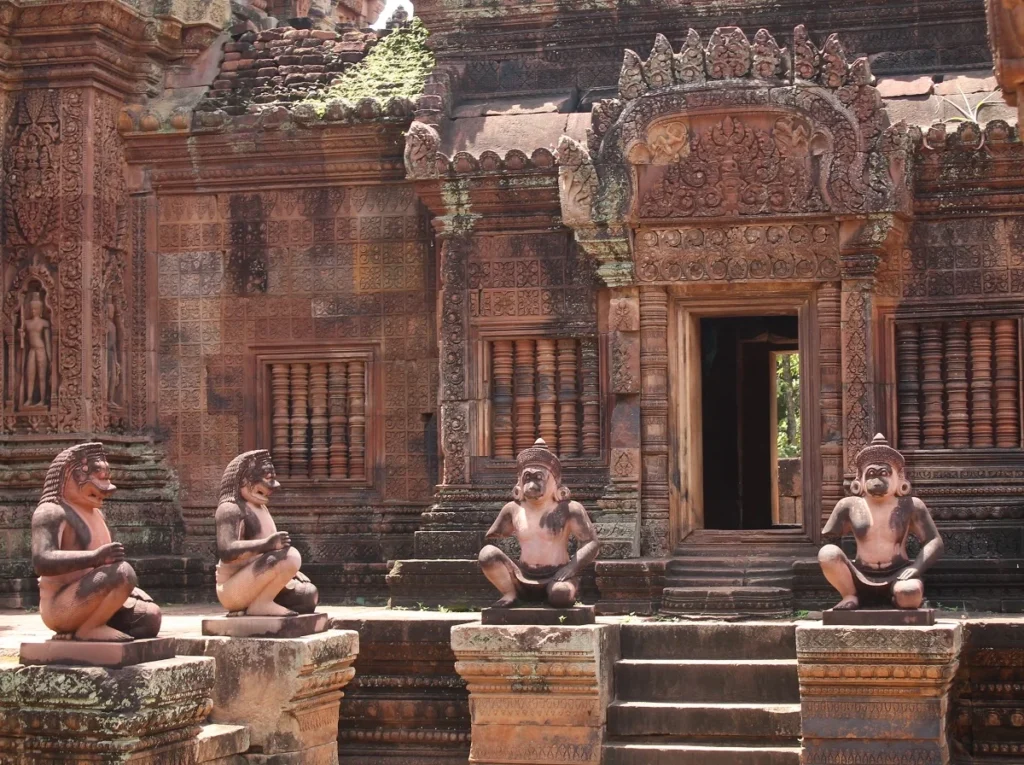 Aufnahme von zwei wachenden Löwen sowie zwei Affen-Skulpturen im Tempel von Banteay Srei. Die Figuren sind in der Hocke und haben menschliche Leiber. Tempel aus rosa Sandstein mit feinen Schnitzereien auf der gesamten Fassade. 