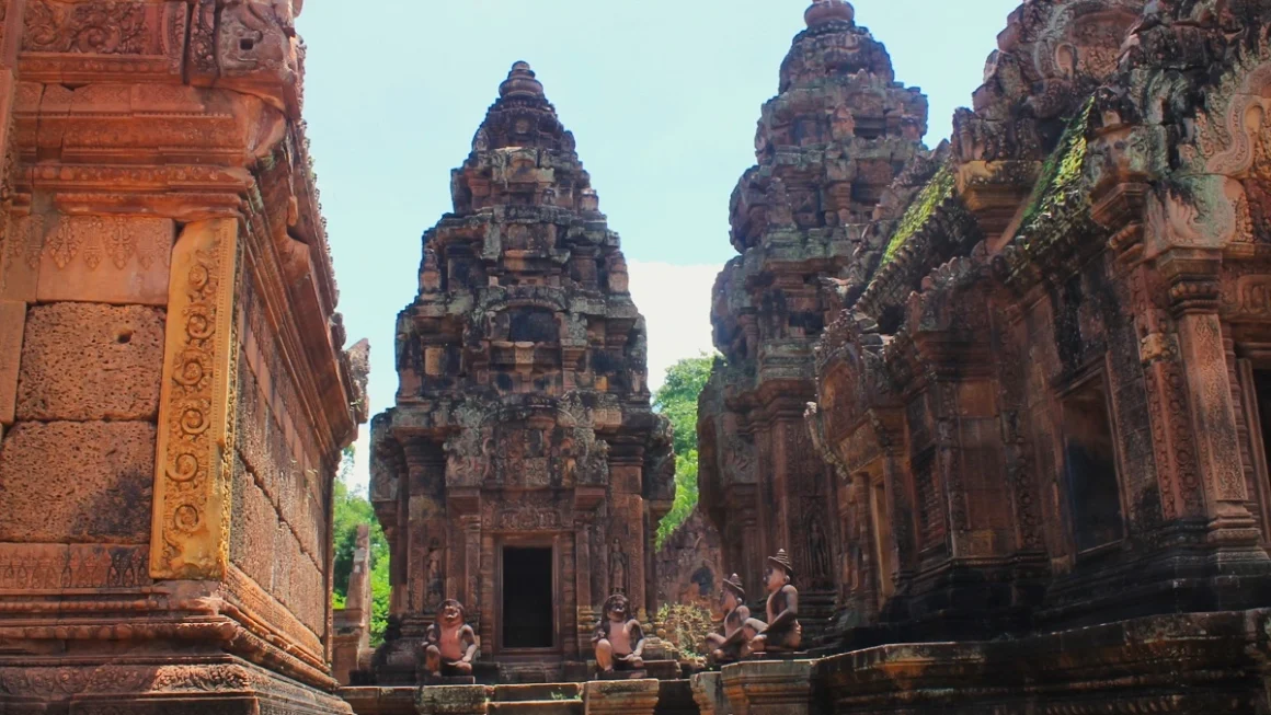 Zentrales Heiligtum von Banteay Srei. Rechts und links Gebäude aus rosa Sandstein und hinten zwei Türme des Tempels. Davor Statuen von hockenden Wachen mit Tierköpfen. Blauer Himmel.