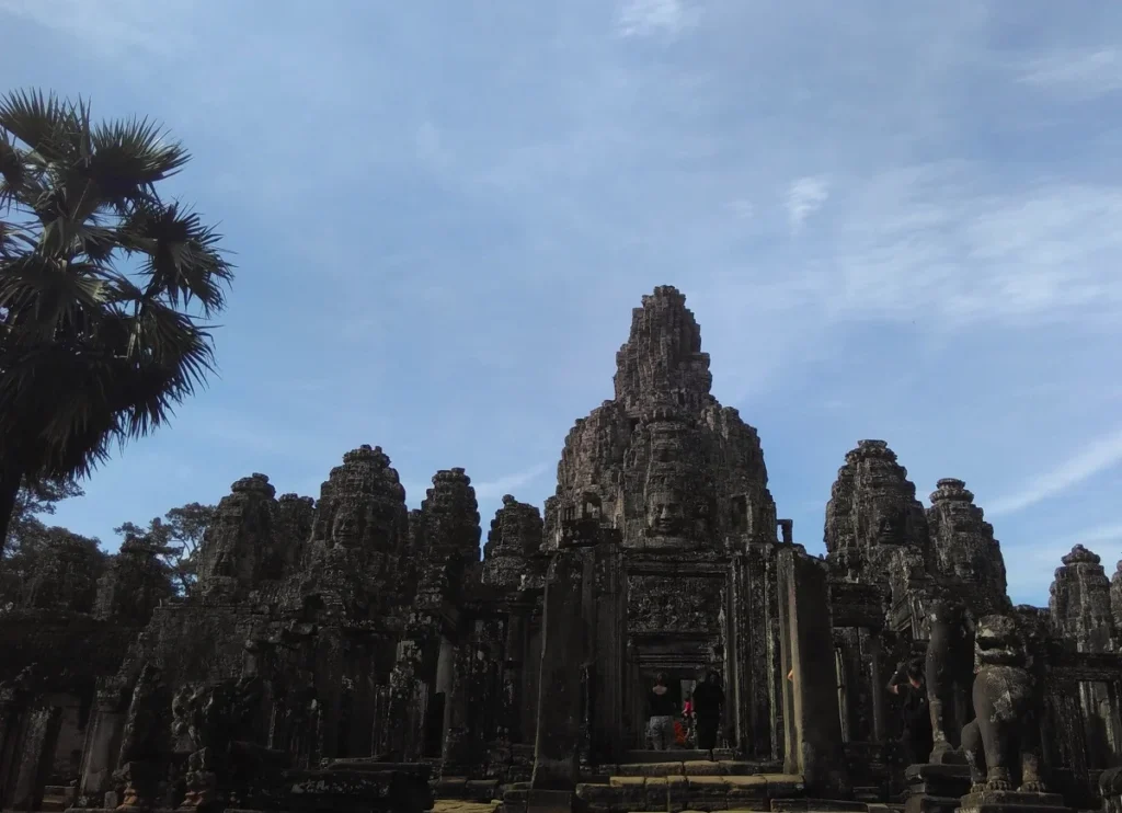 Bayon Tempel mit Türmen und Gesichtern vor blauem Himmel und leicht bewölktem Hintergrund. Eine Palme ragt links ins Bild.