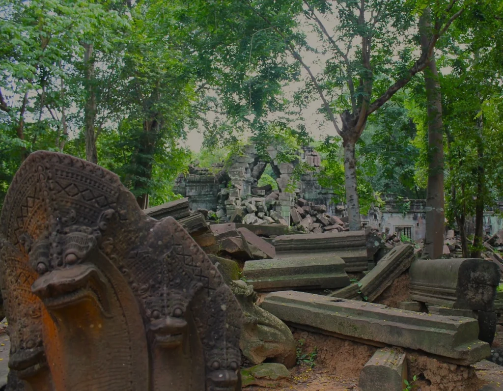 Blick auf Trümmer am Eingang zum Tempel Beng Maelea. Im Vordergrund vier Köpfe der Naga-Schlange. Im Hintergrund grüne Bäume des Dschungels. 