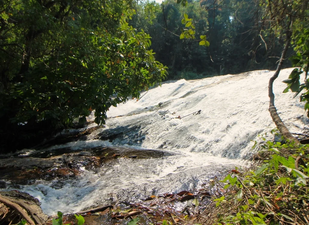 Breiter, flacher Wasserfall auf Felsen am Pha Dok Siew Nature Trail. Von der Seite. Über das rauschende Wasser ragen grüne Bäume.  
