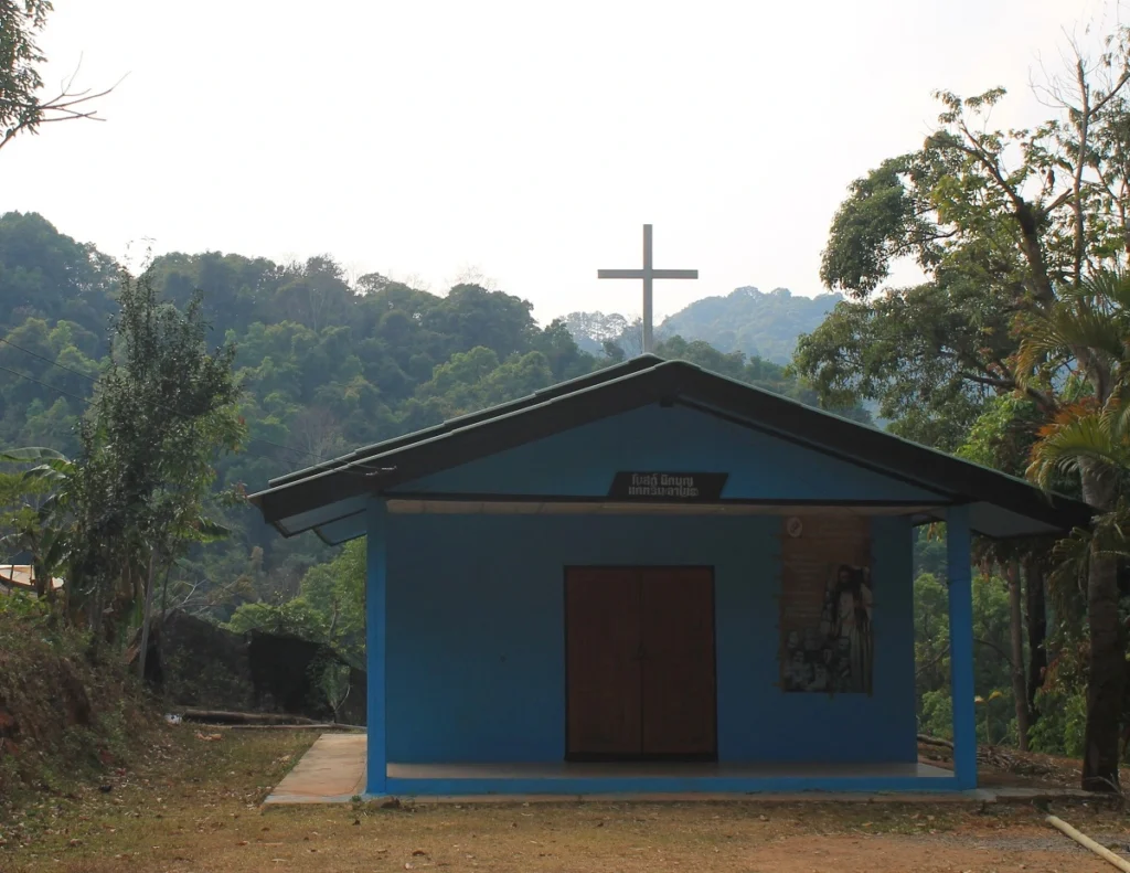 Kirche in Mae Klang Luang. Blau gestrichenes Haus mit Giebel über brauner Holztür. Auf dem Dach ein großes weißes Kreuz. Im Hintergrund die grünen Hügel im Nationalpark Doi Inthanon. 