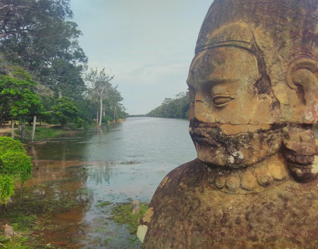 Seitliche Nahaufnahme Büste eines Gottes auf der  Naga-Balustrade am Südtor von angkor Thom. Dahinter Wassergraben mit begrüntem Ufer. 