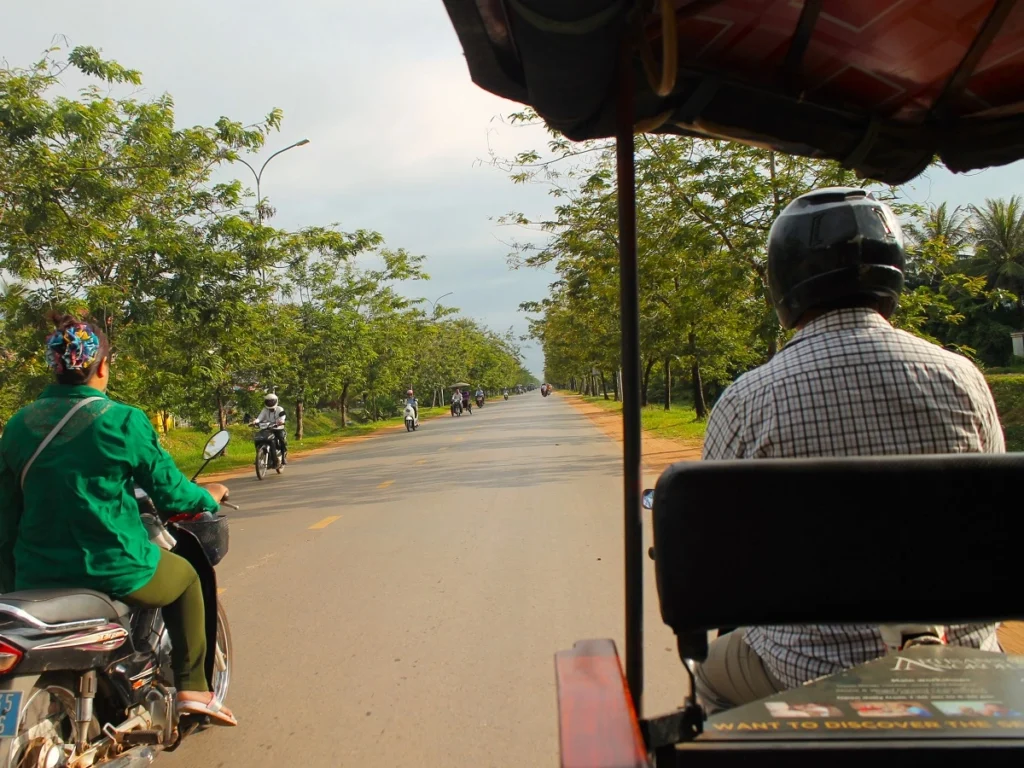 Tuk Tuk auf zweispuriger Straße in Kambodscha. Rechts Tuk Tuk Fahrer mit schwarzem Helm. Links Frau in grünem Hemd auf überholendem Motorrad. Grüne Bäume am Straßenrand. Entgegenkommende Motorbikes und Tuk Tuks. 