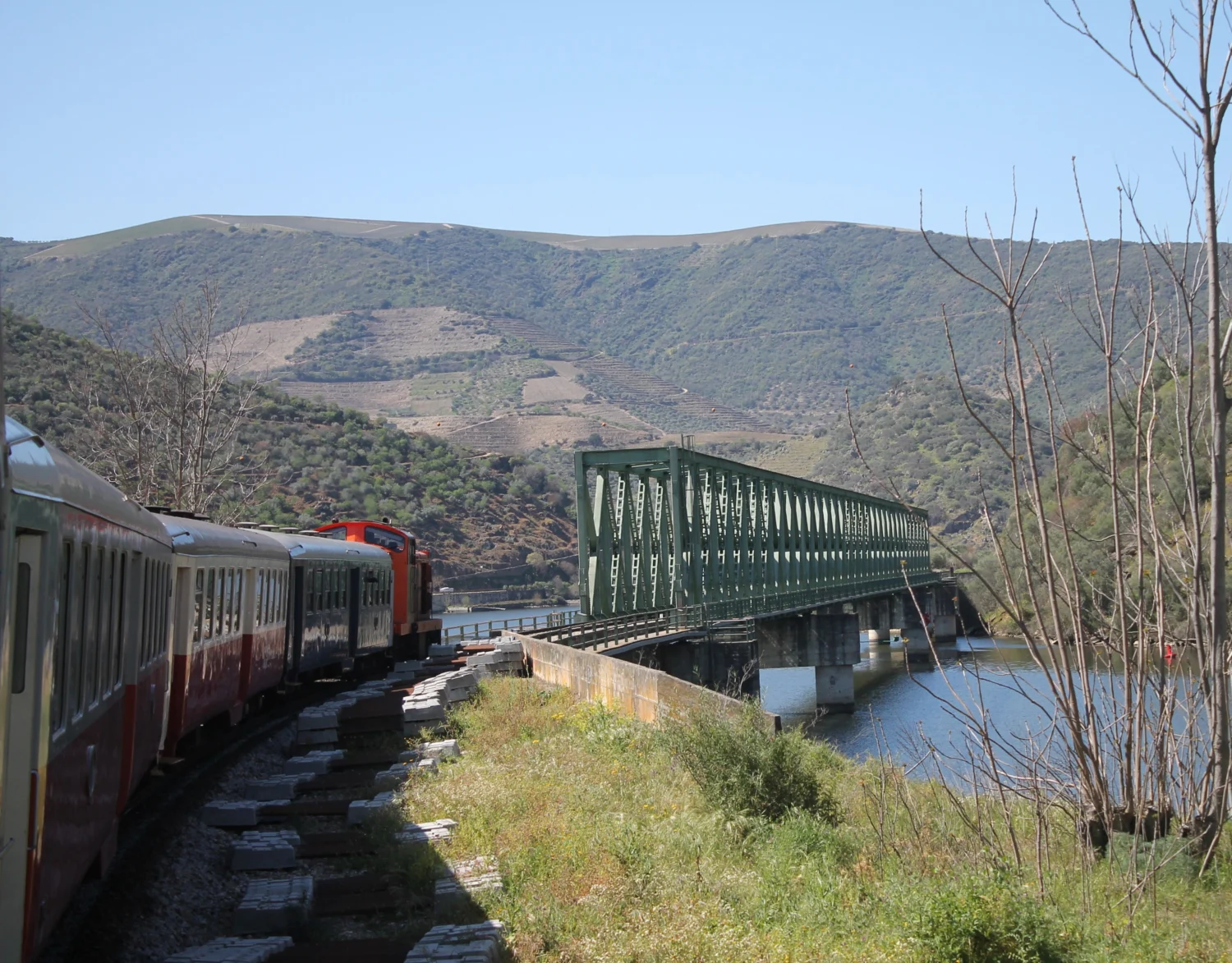 Zug fährt in Rechtskurve über Metallbrücke vor hohem Bergkamm. Blick aus dem Zugfenster