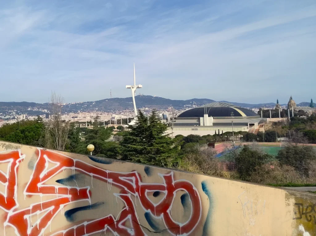 Blick über eine Mauer mit Graffiti-Schriftzug über den Olympischen Park in Barcelona. Vorne der Fernsehturm und der Palau Sant Jordi. Hinten der Berg Tibidabo mit dem Torre de Collserola.