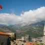 Fahne von Montenegro auf Zitadelle Budva, dahinter Kirchturm der St.-Ivan-Kirche. Begrünte Karstberge mit Wolken im Hintergrund.