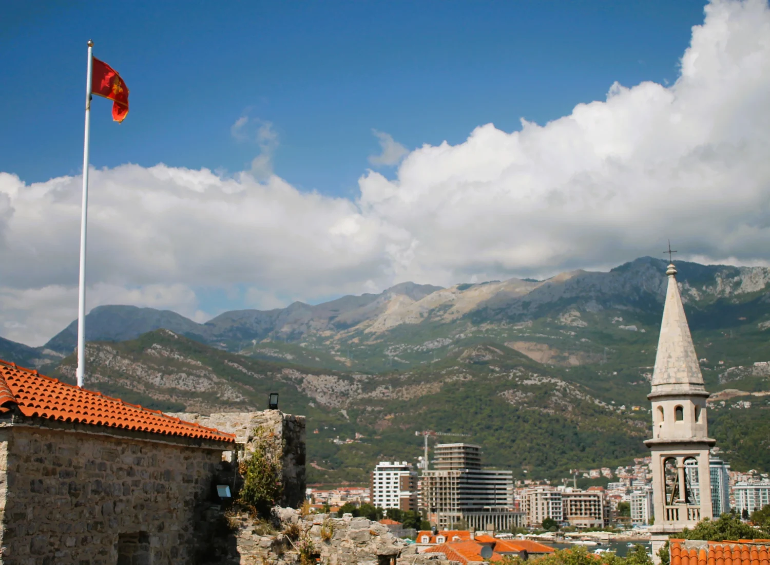 Fahne von Montenegro auf Zitadelle Budva, dahinter Kirchturm der St.-Ivan-Kirche. Begrünte Karstberge mit Wolken im Hintergrund.