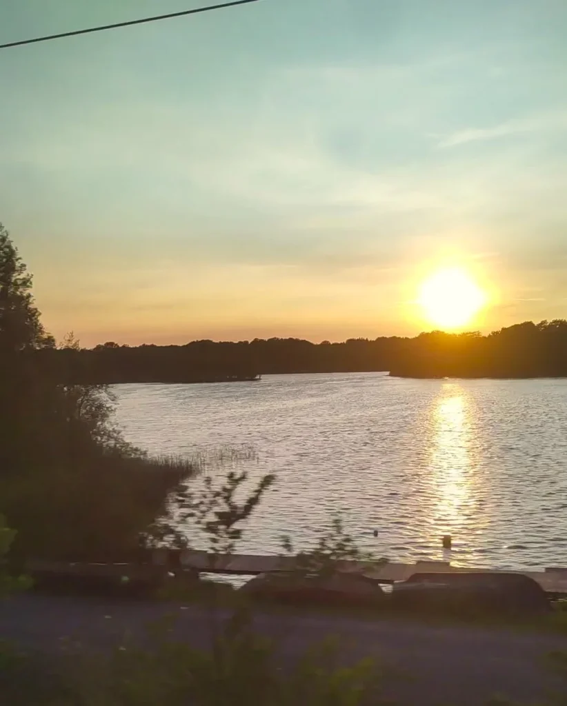 Überbelichtete, untergehende Sonne hinter einem See mit Wald. 