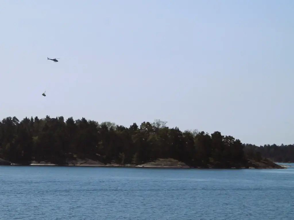 Hubschrauber transportiert großes Paket an einem langen Seil über eine Schäreninsel. 