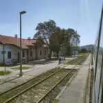 Kroatien mit dem Zug - am Bahnsteig vom Bahnhof Unesic. Aus dem Zugfenster. Schaffner in Uniform steht vor dem Empfangsgebäude.