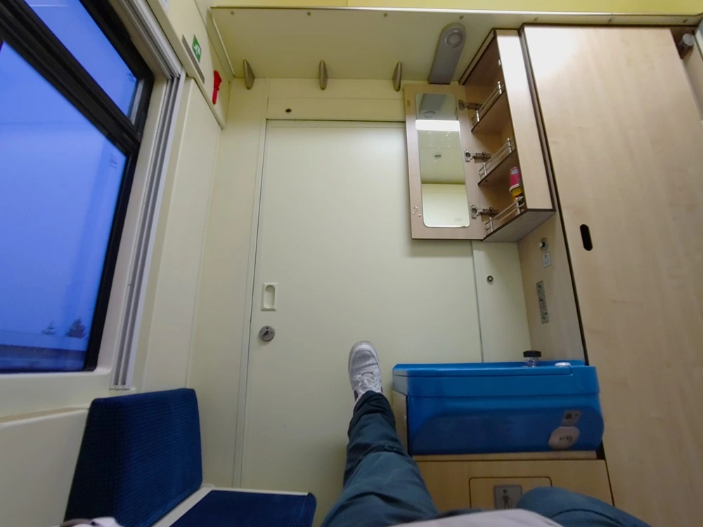Schlafabteil im Euronight 406 vom Bett aus, mit ausgestrecktem Bein zum Waschbecken. Mit Spiegelschrank, Kleiderschrank, Gepäckablage, Extra-Sitzgelegenheit und Fenster. East Rail Stories