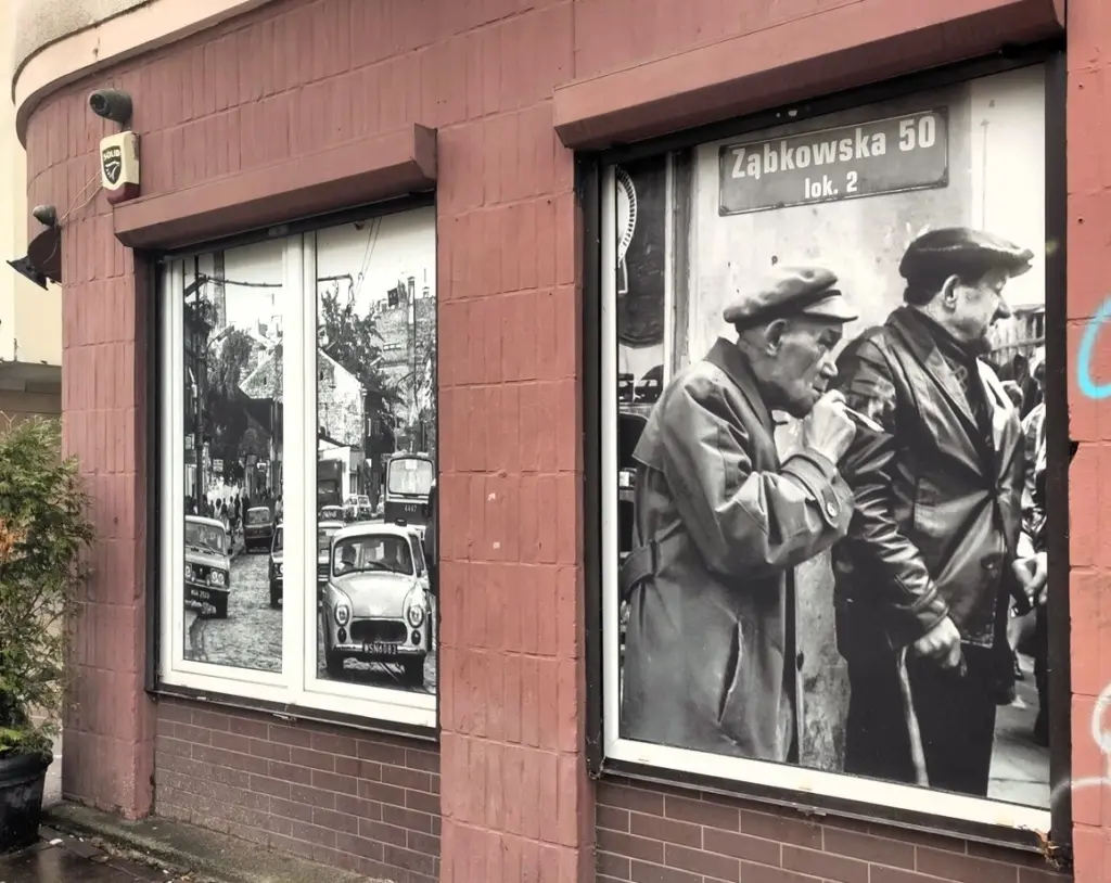 Zabkowska 50 - Schwarzweiß-Foto am Fenster zeigt alte Autos auf der Straße und ältere Männer mit Schiebemützen - vermutlich Arbeiter. 
East Rail Stories
