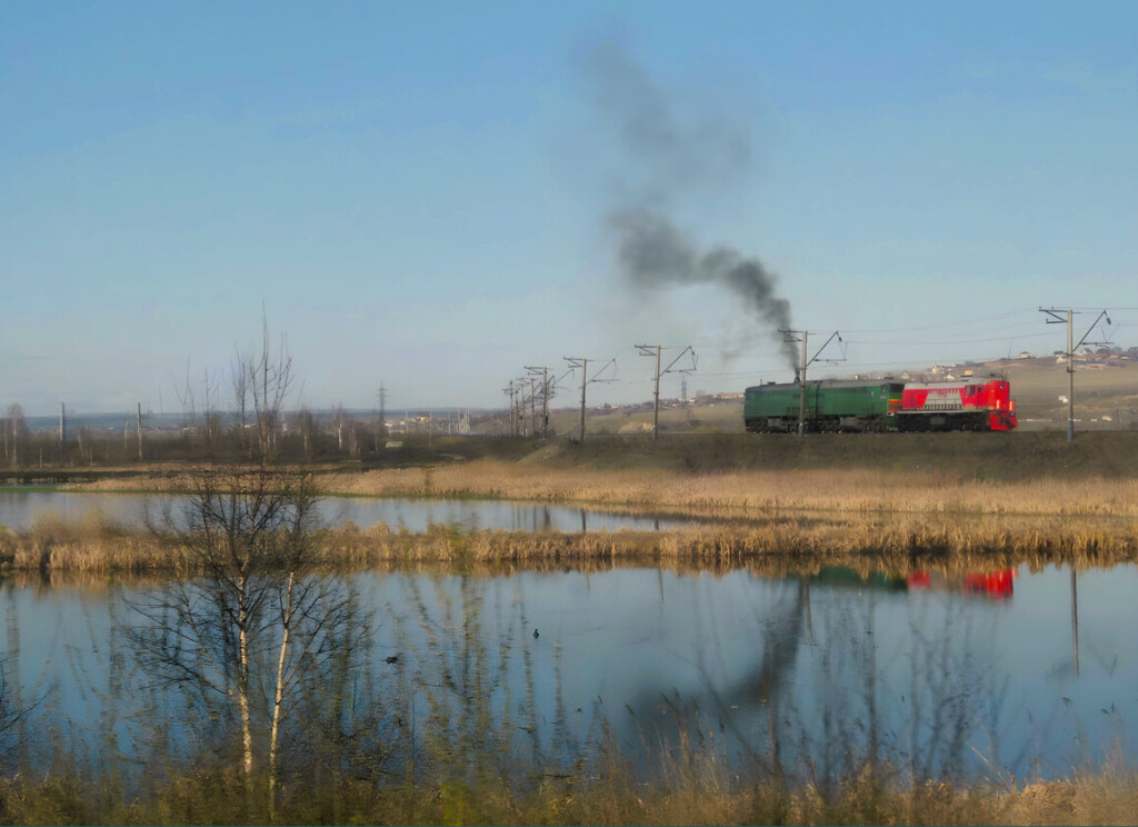 Russische Lokomotive stößt schwarzen Rauch in die Luft. Spiegelung in einem See.
East Rail Stories
