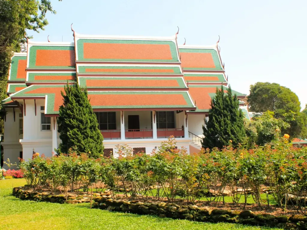 Seitenansicht Bhubing Palast. Dachbedeckung helles Orange mit grüner Umrandung. Davor grüner Garten. 