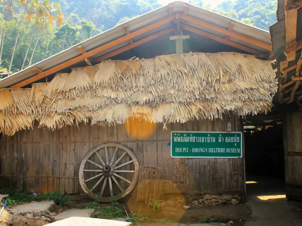 Holz- und Strohhütte des Hmong Hilltribe Museum in Doi Pui. An der Hütte lehnt ein Kutschenrad. 