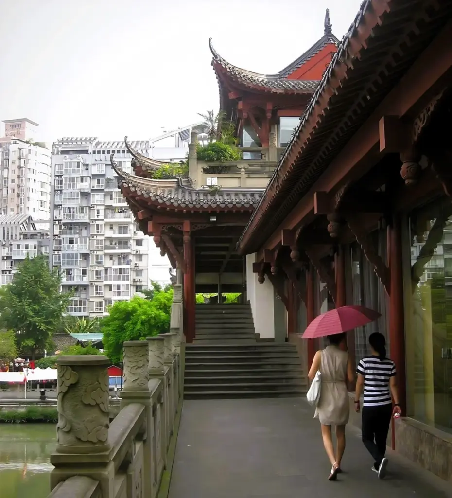 Zwei Frauen laufen über die Anshun Brücke in Chengdu. Eine Frau trägt einen rosa Regenschirm. Das steinerne Brückengeländer trägt kleine ornamentierte, runde Säulen.  