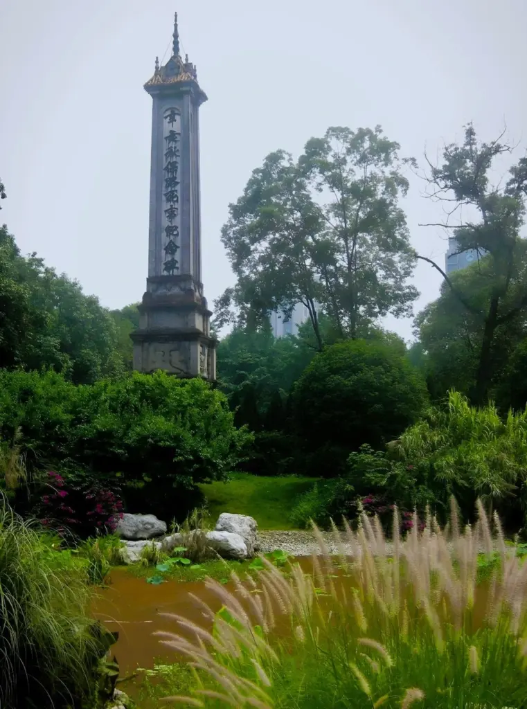Das Monument der Märtyrer der Eisenbahnschutzbewegung im Volkspark von Chengdu ragt zwischen den grünen Büschen und Bäumen hervor. Im Vordergrund ein kleiner Teich mit Schilf-Bepflanzung. 