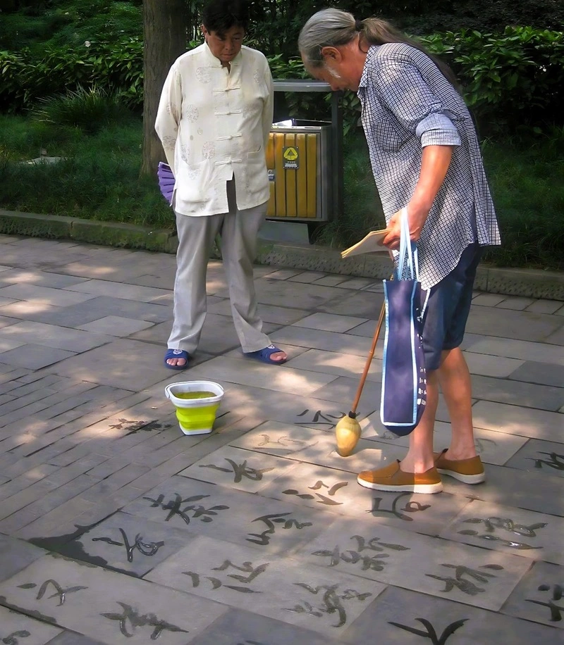 Ein Dishu-Künstler zeichnet mit einem großen Pinsel und Wasser chinesische Schriftzeichen auf den Betonboden. Ein Mann sieht ihm zu.