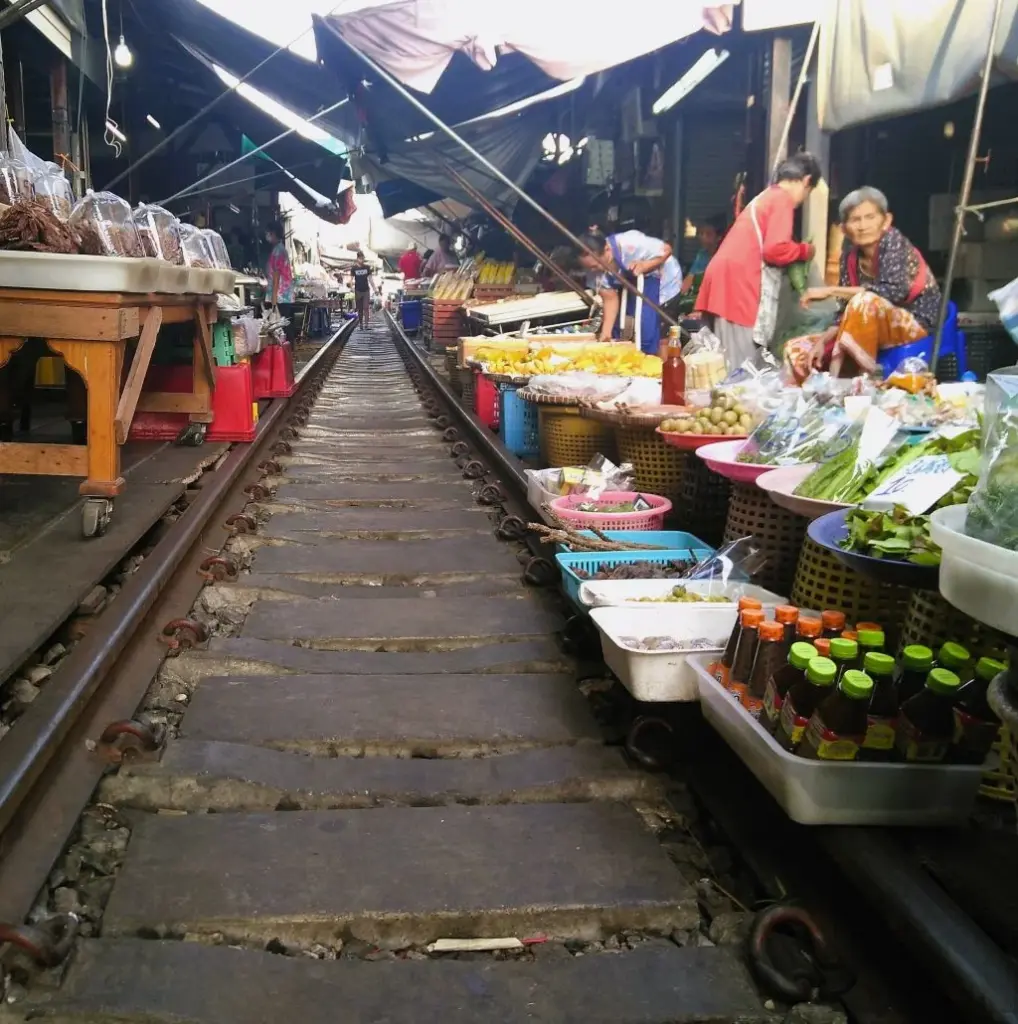 Gleis im leeren Mae Klong Markt. Rechts uns links Verkaufsstände und Verkäuferinnen. Über den Markt ragen Sonnenschirme. 