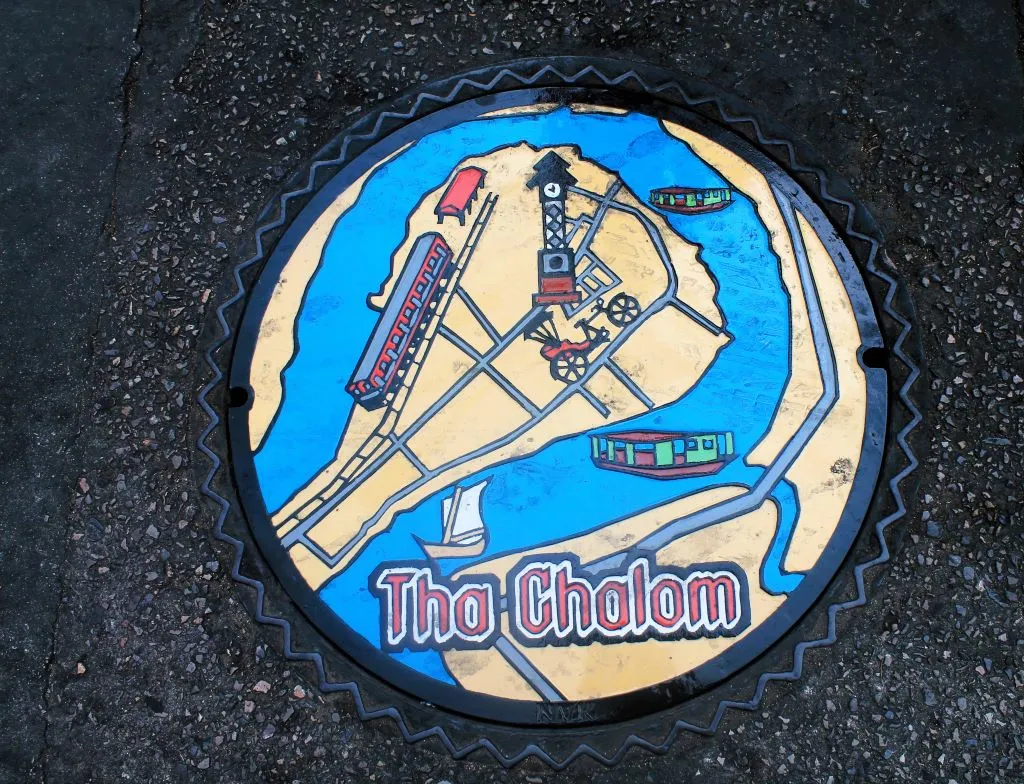 Gullydeckel auf Straße in Tha Chalom mit künstlerisch gestalteter Landkarte des Ortes. Auf der Karte sind Züge. Fähren und ein eine Rikscha eingezeichnet. 