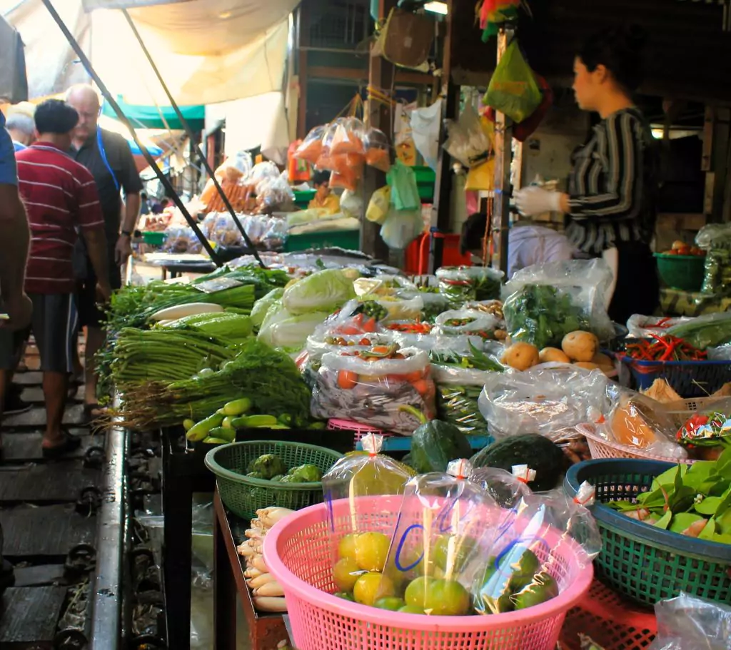 Gemüsestand auf dem Mae Klong Railway Market mit einigen westlichen Besuchern auf den Schienen. 