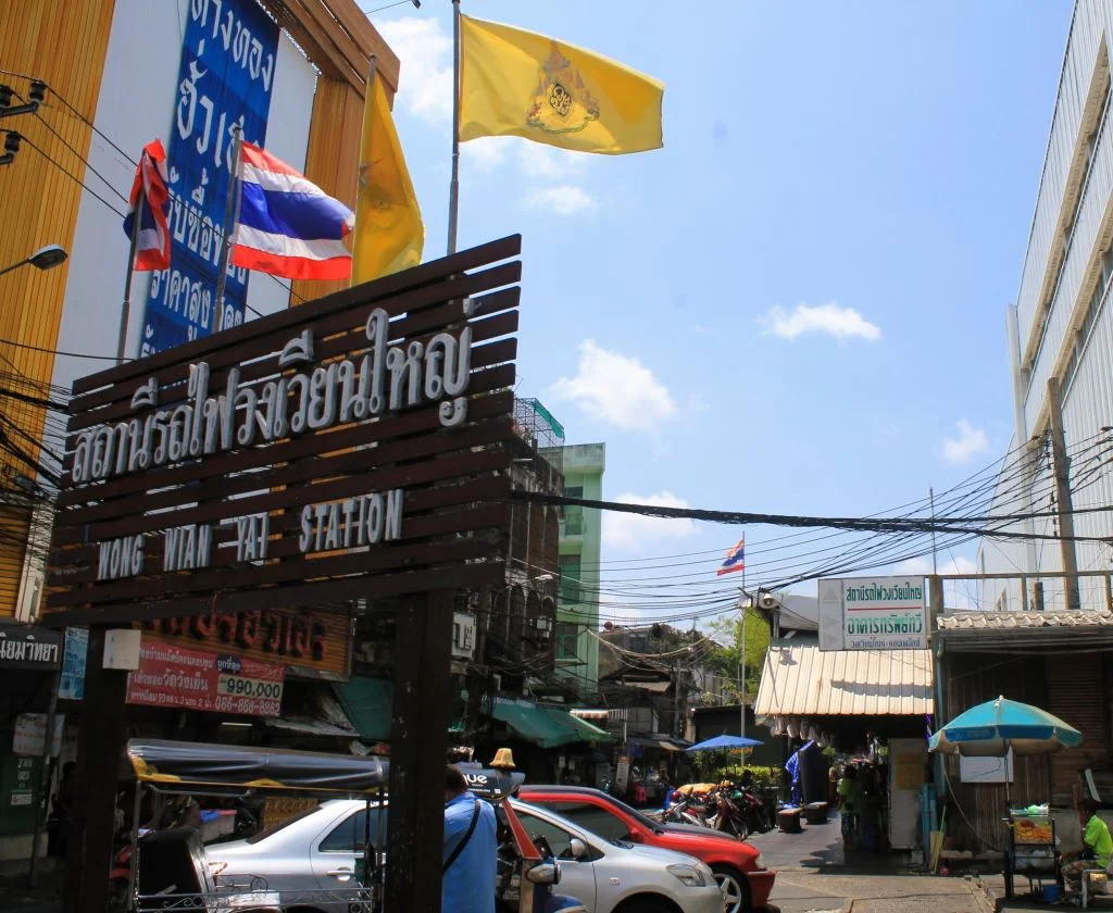 Eingang zum Bahnhof Wong Wian Yai in Bangkok. Schild aus braunen Holzlatten mit weißer Aufschrift in Thai und Englisch. Darauf die Fahnen Thailands und des Königshauses. 