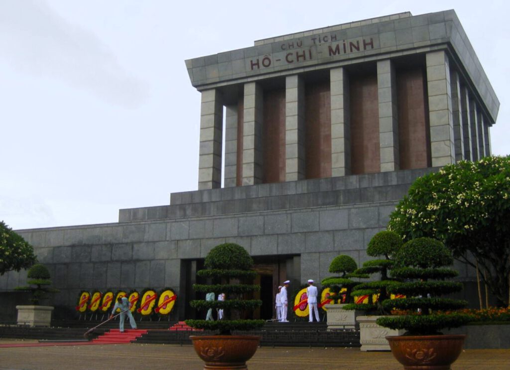 Ho Chi Minh Mausoleum (Chu Tich) in Hanoi. Rechteckiger Betonbau in Nahaufnahme. Davor weiß gekleidete Wachen und Ehrenkränze. 