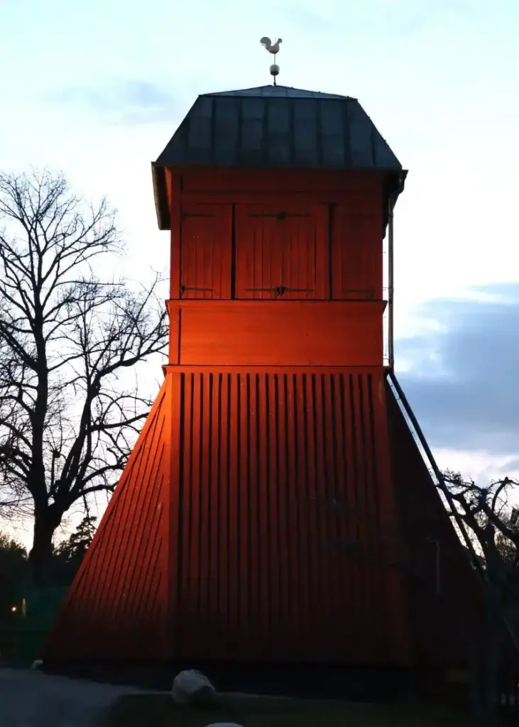Nahaufnahme des Klockstapels von Vaxholm, ein historischer Glockenturm aus rotem Holz. Auf der Spitze sitzt ein Wetterhan. 