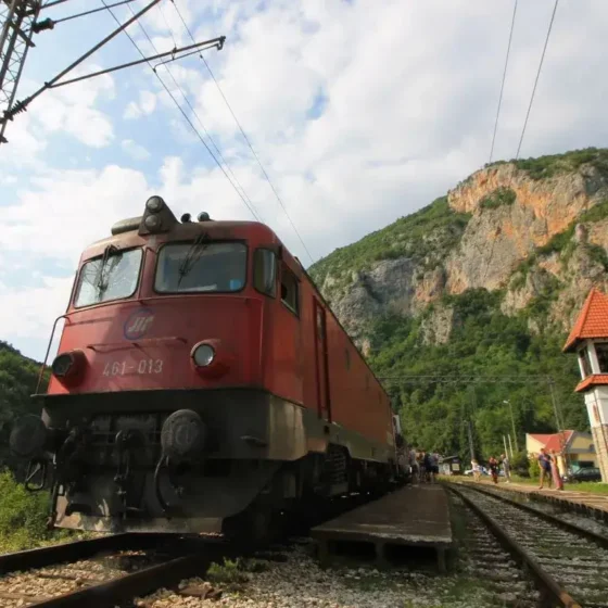 Frontansicht rote Lokomotive der serbischen Eisenbahn am Bahnhof von Vrbnica, nahe der Grenze Serbien-Montenegro. Menschen am Bahnsteig. Hintergrund Karsthügel.