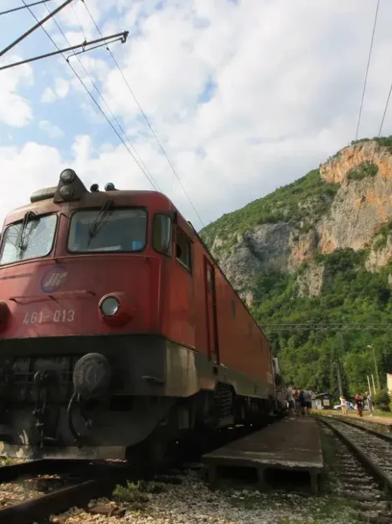 Frontansicht rote Lokomotive der serbischen Eisenbahn am Bahnhof von Vrbnica, nahe der Grenze Serbien-Montenegro. Menschen am Bahnsteig. Hintergrund Karsthügel.