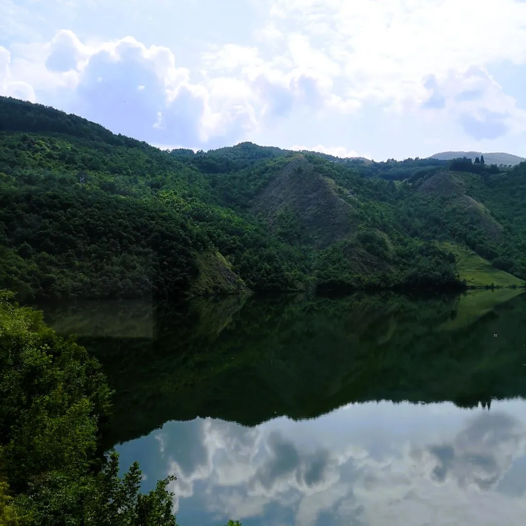 Grüne Hügellandschaft im Norden von Montenegro. Bewaldete Berge spiegeln sich in einem See.