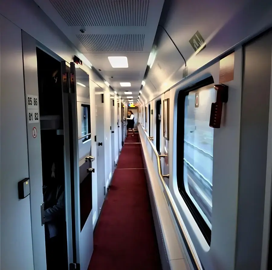 Nachtzug Berlin-Moskau. Im Inneren des Schlafwagens ein roter Teppich auf dem Boden. Links offene Türen zu den Schlafabteilen. Rechts die Fensterzeile. Im Hintergrund eine Schaffnerin.  