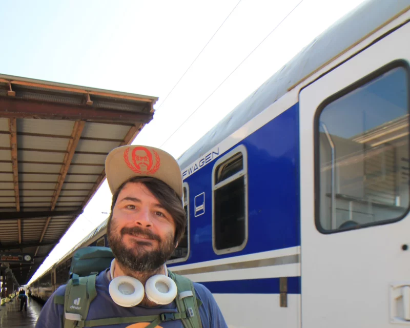 Meine Geschichte - Von kurzen Osteuropa Trips zu ausufernden East Rail Stories. Ich stehe mit Kopfhörern um den Hals vor einem blau-weißen Schlafwagen am Bahnsteig.