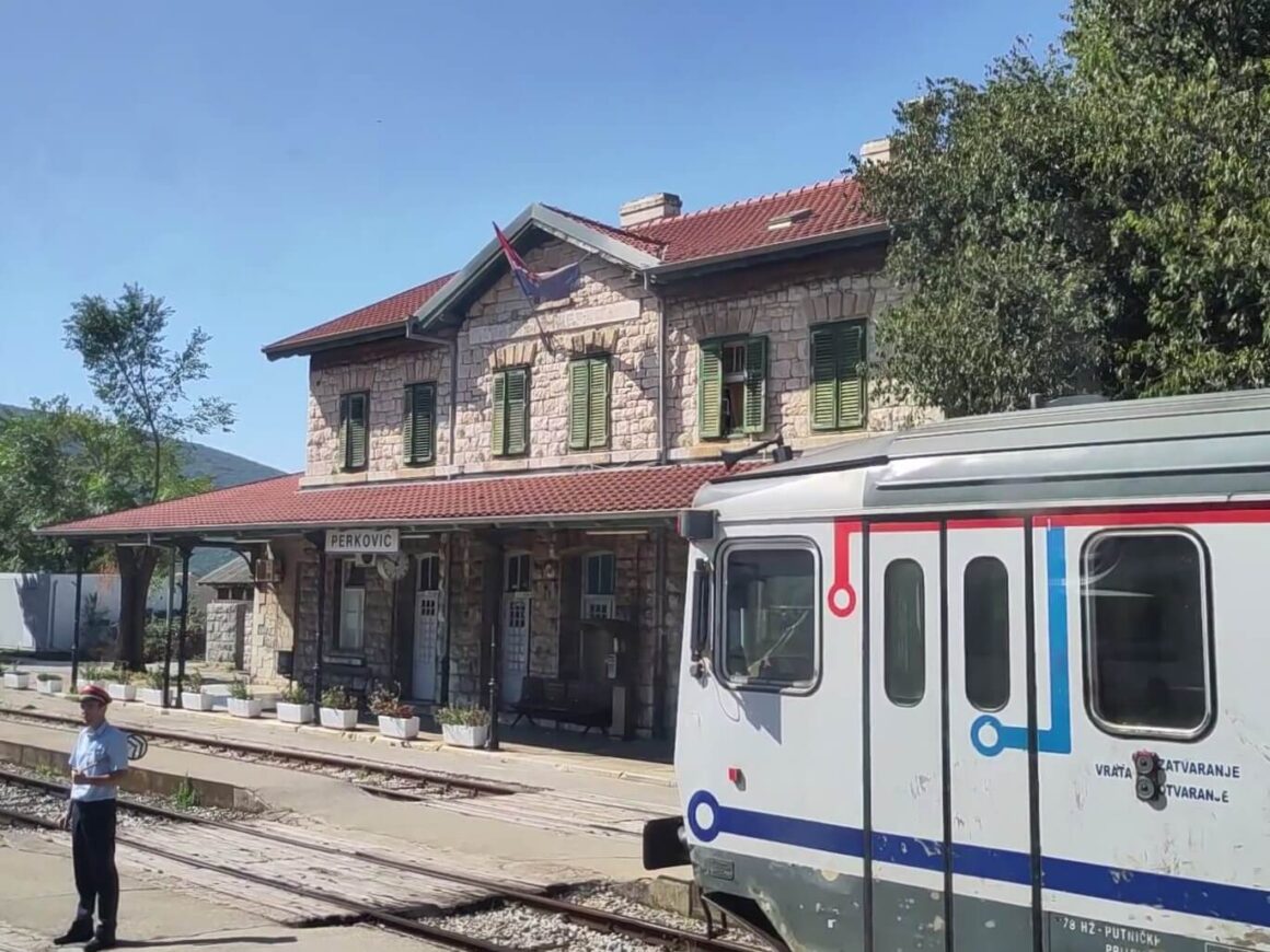 Eein Eisenbahner mit Kelle steht neben anderem Zug, vor dem schönem steinernen Bahnhofsgebäude in Perković. 
East Rail Stories
