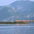 Zug fährt über eine Eisenbahnbrücke in Virpazar Montenegro. Orangefarbene Lokomotive. Dahinter grüner Berge. Perspektive vom Skadarsee aus.