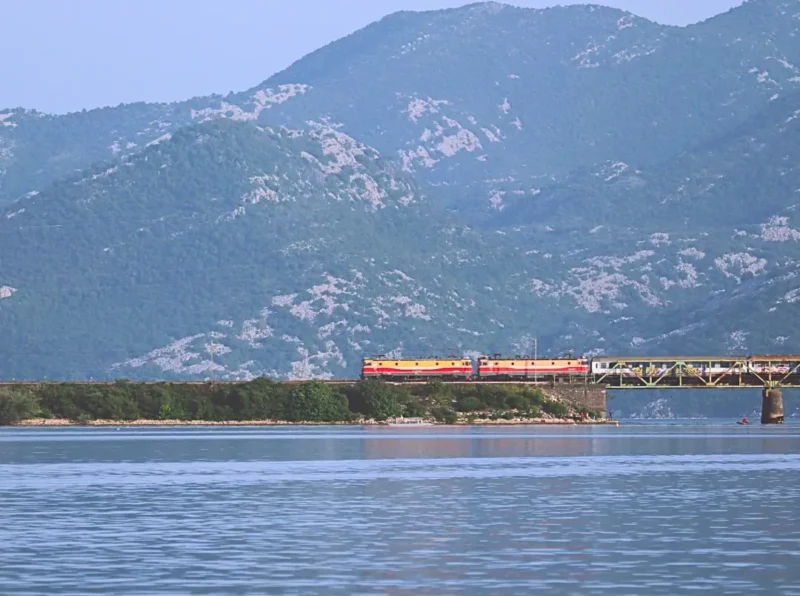Zug fährt über eine Eisenbahnbrücke in Virpazar Montenegro. Orangefarbene Lokomotive. Dahinter grüner Berge. Perspektive vom Skadarsee aus.
