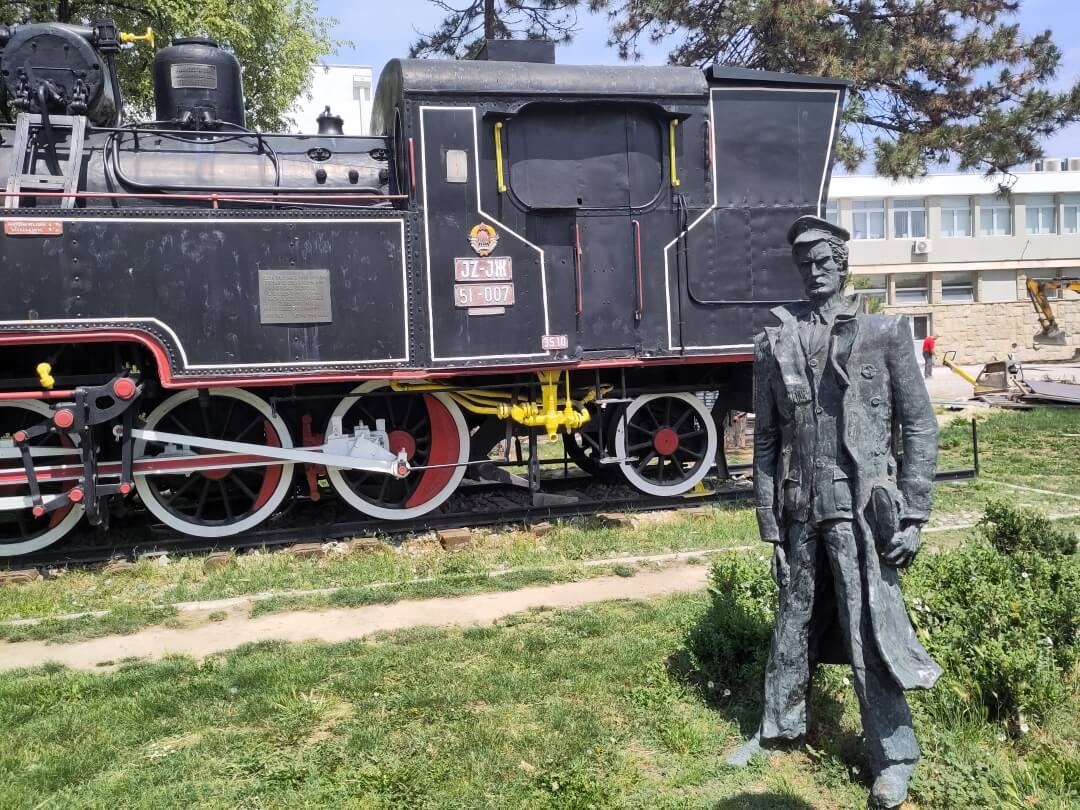 Alte Lokomotive von 1925 ist auf einer Wiese ausgestellt. Davor die Bronzefigur eines Eisenbahnarbeiters. 