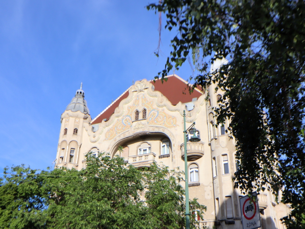 Großes Jugendstilgebäude mit heller Fassade und rotem Dach hinter grünen Bäumen.