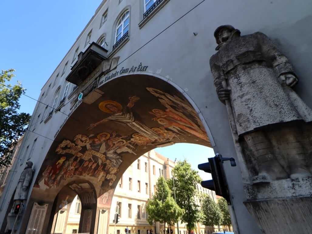 Betonfassade eines Torbogens mit Soldatenreliefs. Der Bogen ist mit christlichen Fresken bemalt.