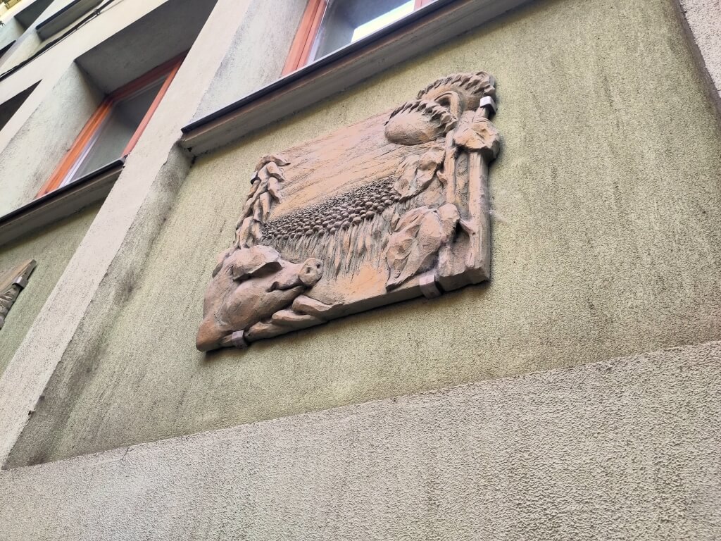 Relief-Kachel an Haussfassade. Ein Schwein blickt auf ein Feld von Sonnenblumen.