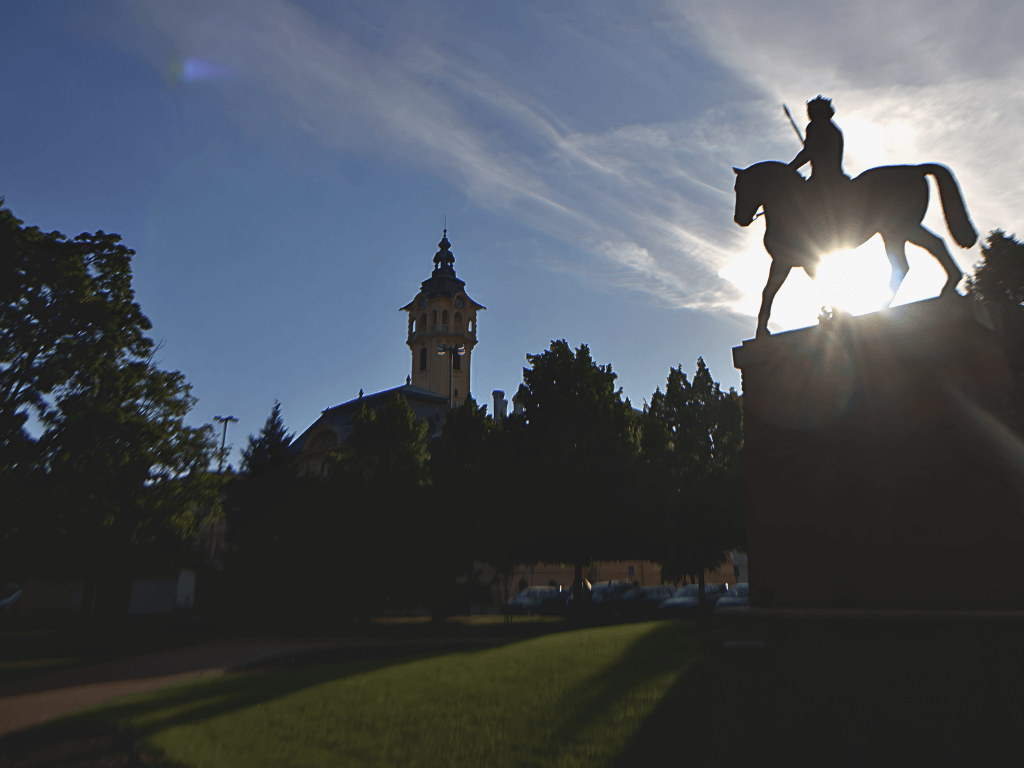 Park bei mit Reiterstatue, gegen die Sonne fotografiert, mit der Silhouette des Szegeder Rathauses im Hintegrund.