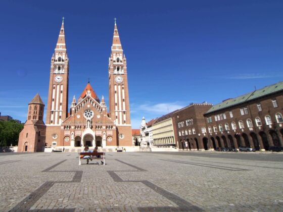 Zwei Personen sitzen auf Parkbank mitten auf Domplatz in Szeged vor der Votivkirche,